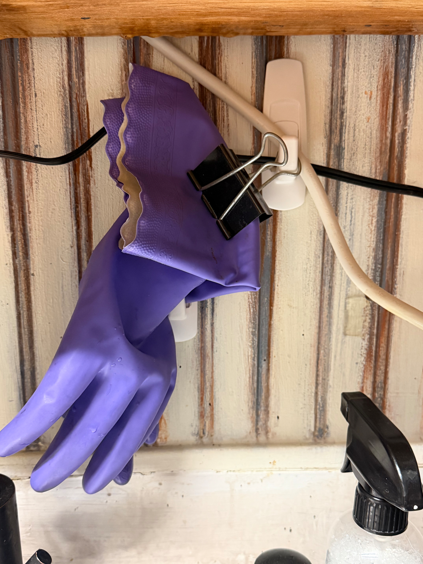 A pair of purple rubber gloves clipped to a command hook with a binder clip, hanging above a spray bottle and an electrical cord.
