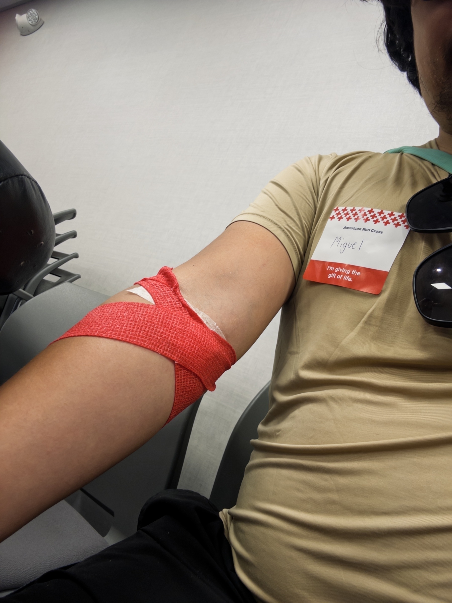 Person in tan shirt after donating blood, arm extended on donation chair with red gauze cuff and white cotton. American Red Cross name tag reads “Miguel” with “I’m giving the gift of life” message.​​​​​​​​​​​​​​​​