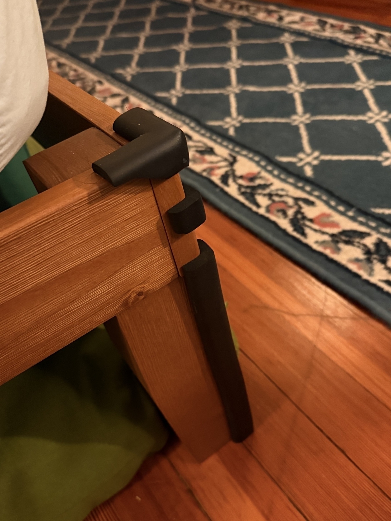 A wooden bed frame corner with protective black padding is positioned on a hardwood floor next to a patterned rug.