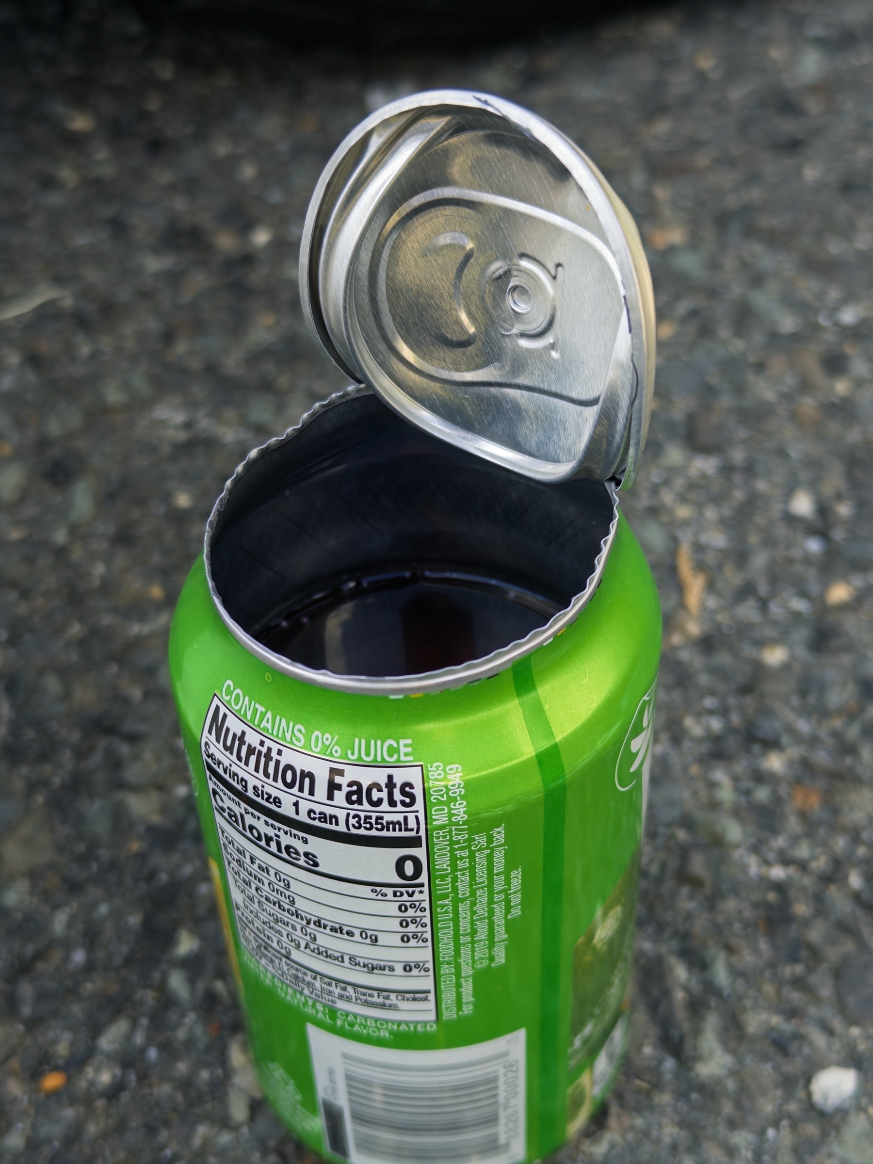 A green soda can with the lid removed is filled with a dark liquid and placed on a textured surface.