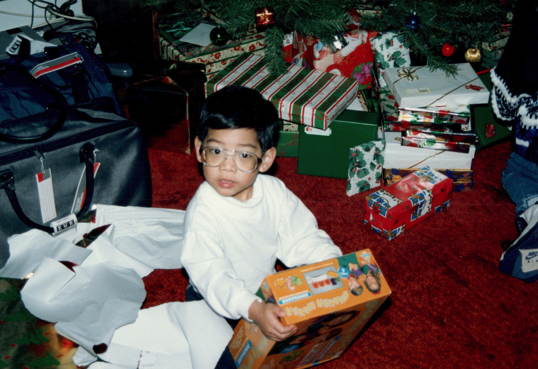 Young boy in white pajamas and glasses sits cross-legged on red carpet beneath Christmas tree, holding orange toy box. Colorfully wrapped presents surround him, creating festive holiday morning scene with cozy, excited atmosphere.​​​​​​​​​​​​​​​​