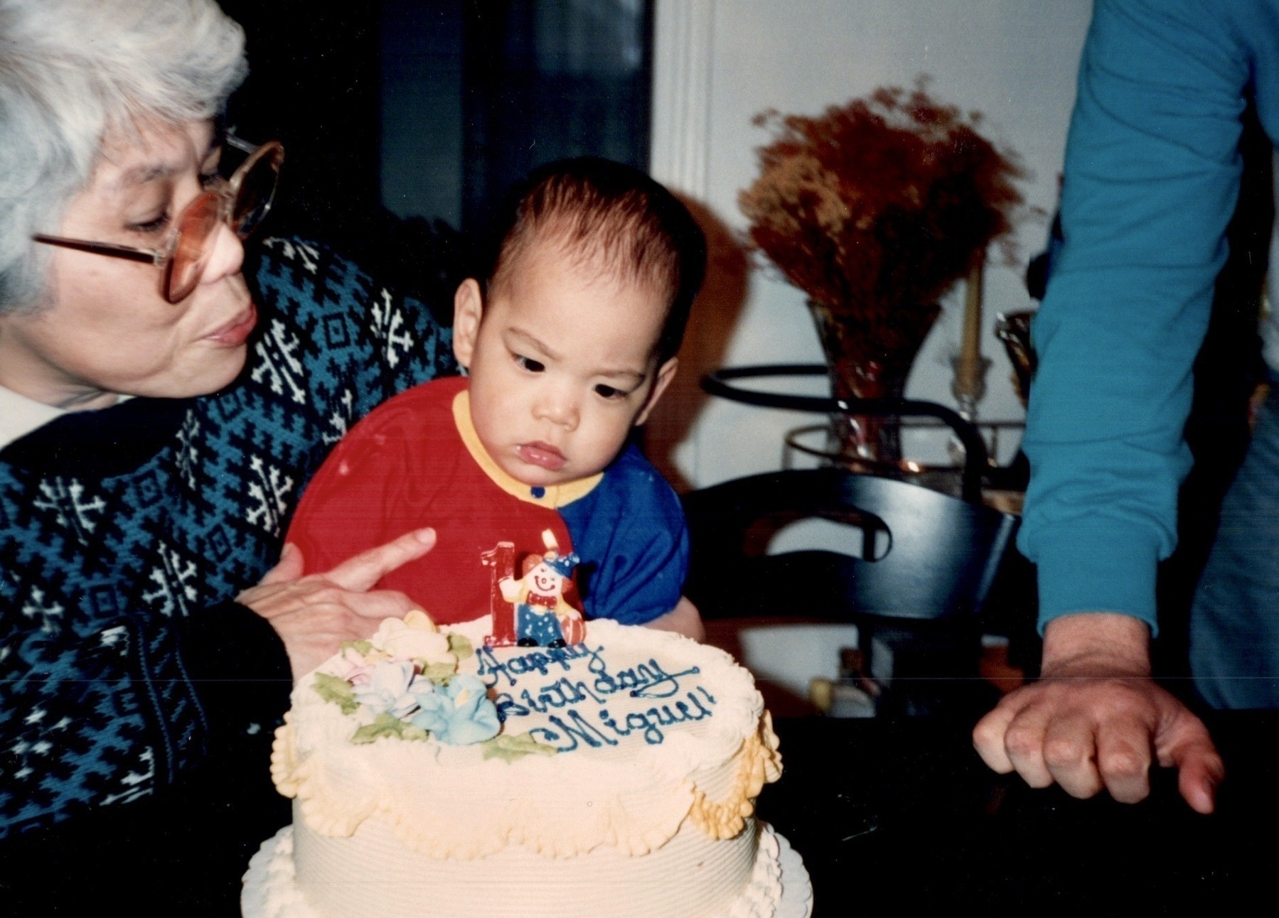 Grandmother with silver hair and glasses holds serious-faced baby in red and blue outfit beside white birthday cake decorated with clown figure. Man in blue shirt stands nearby in warm indoor setting.