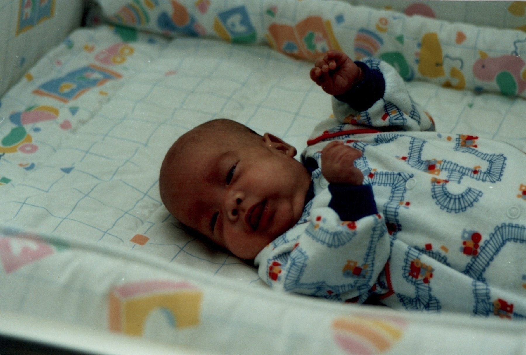 Smiling baby lies on colorful patterned blanket, wearing white onesie with blue trim and small floral details, one fist raised playfully. Soft pastel toys and cheerful nursery patterns fill the cozy background.