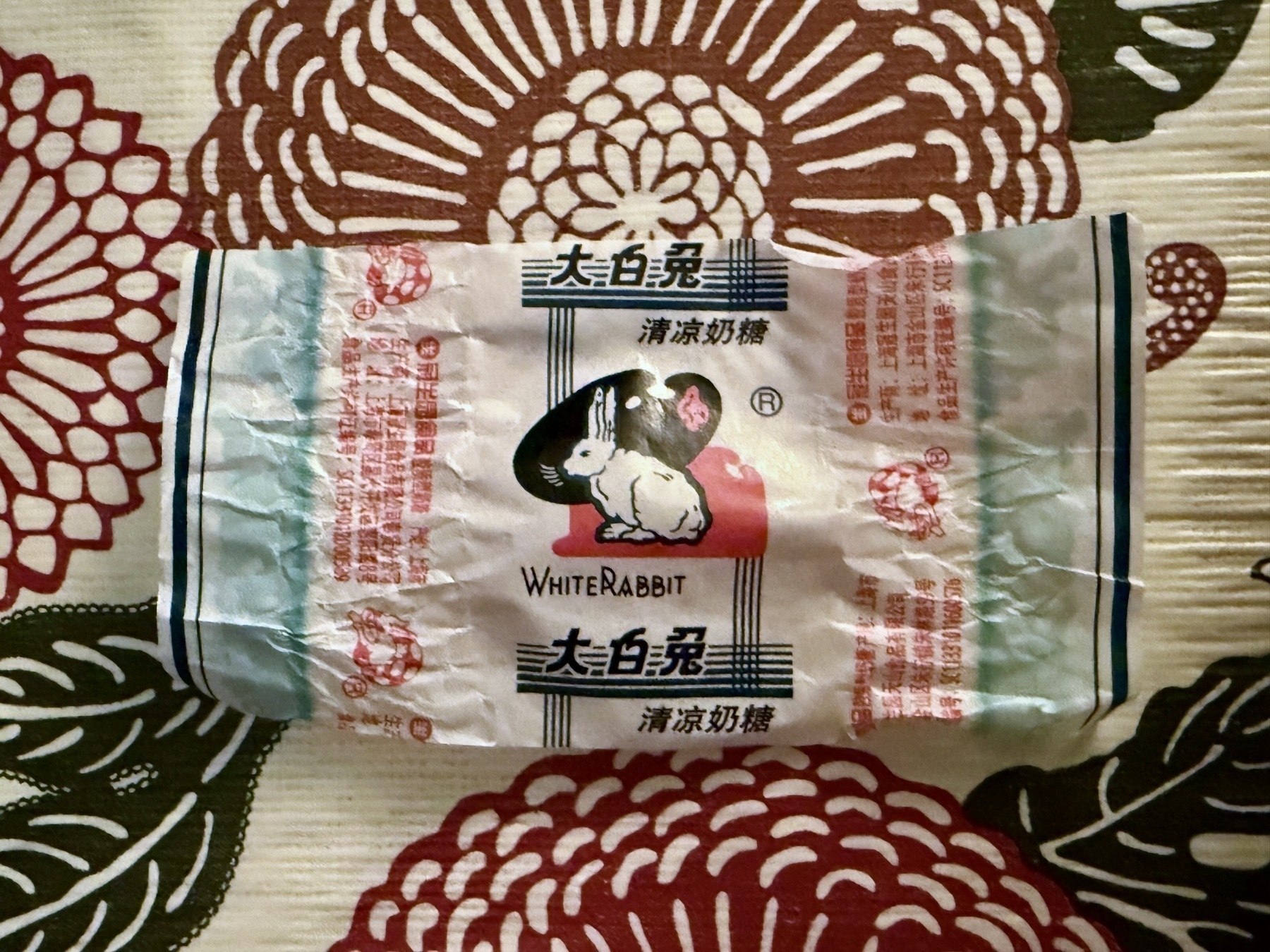 Crumpled white rabbit candy wrapper featuring cartoon character bunny, surrounded by chinese text and blue borders, lying on floral-patterned fabric with burgundy chrysanthemums and cream background