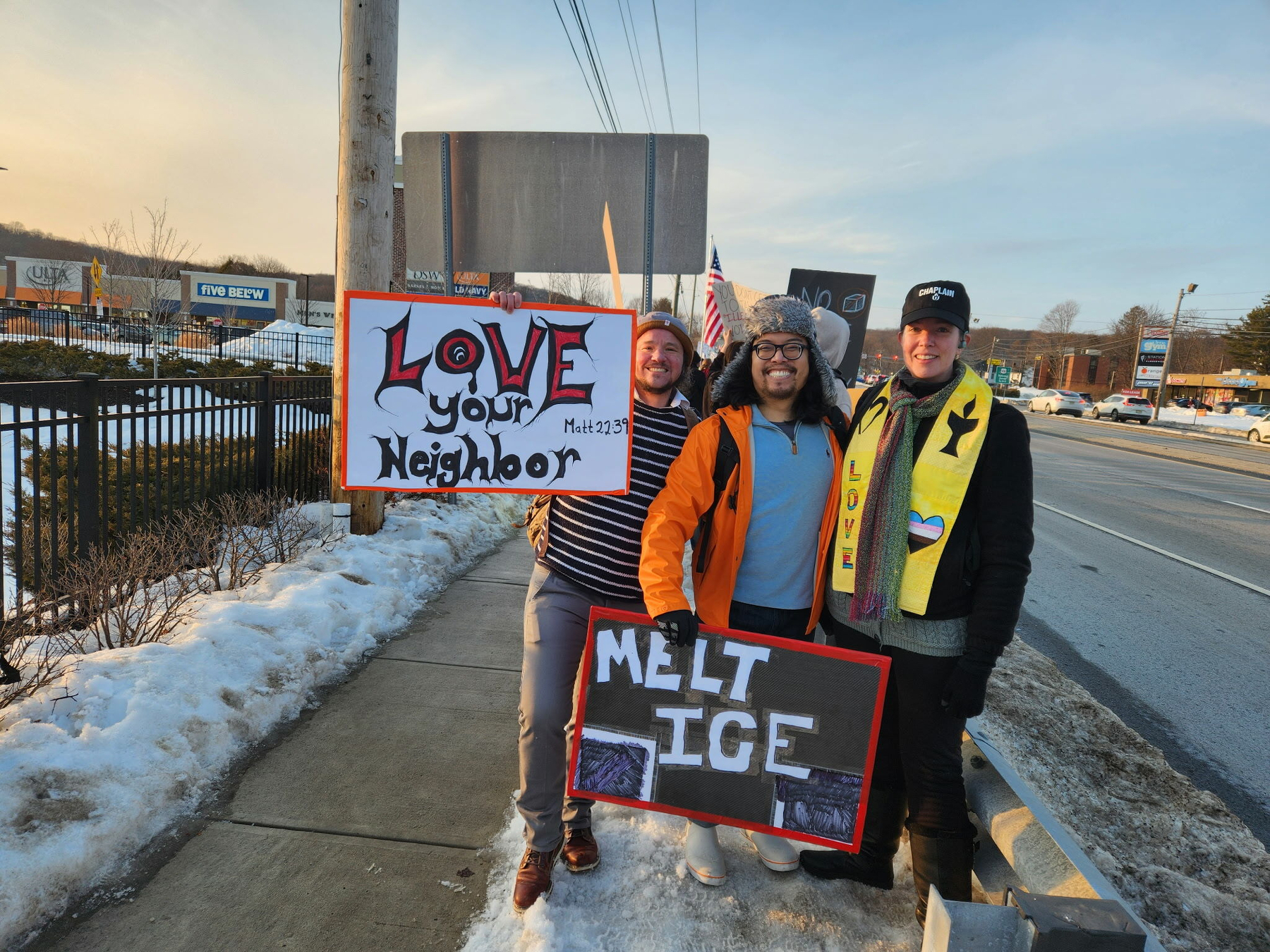 Three smiling protesters stand on a snowy sidewalk holding signs reading “Love Your Neighbor” and “Melt ICE.” One wears a chaplain hat and yellow stole with a trans heart; another an orange jacket; another a striped shirt. Sunset light, suburban backdrop.​​​​​​​​​​​​​​​​