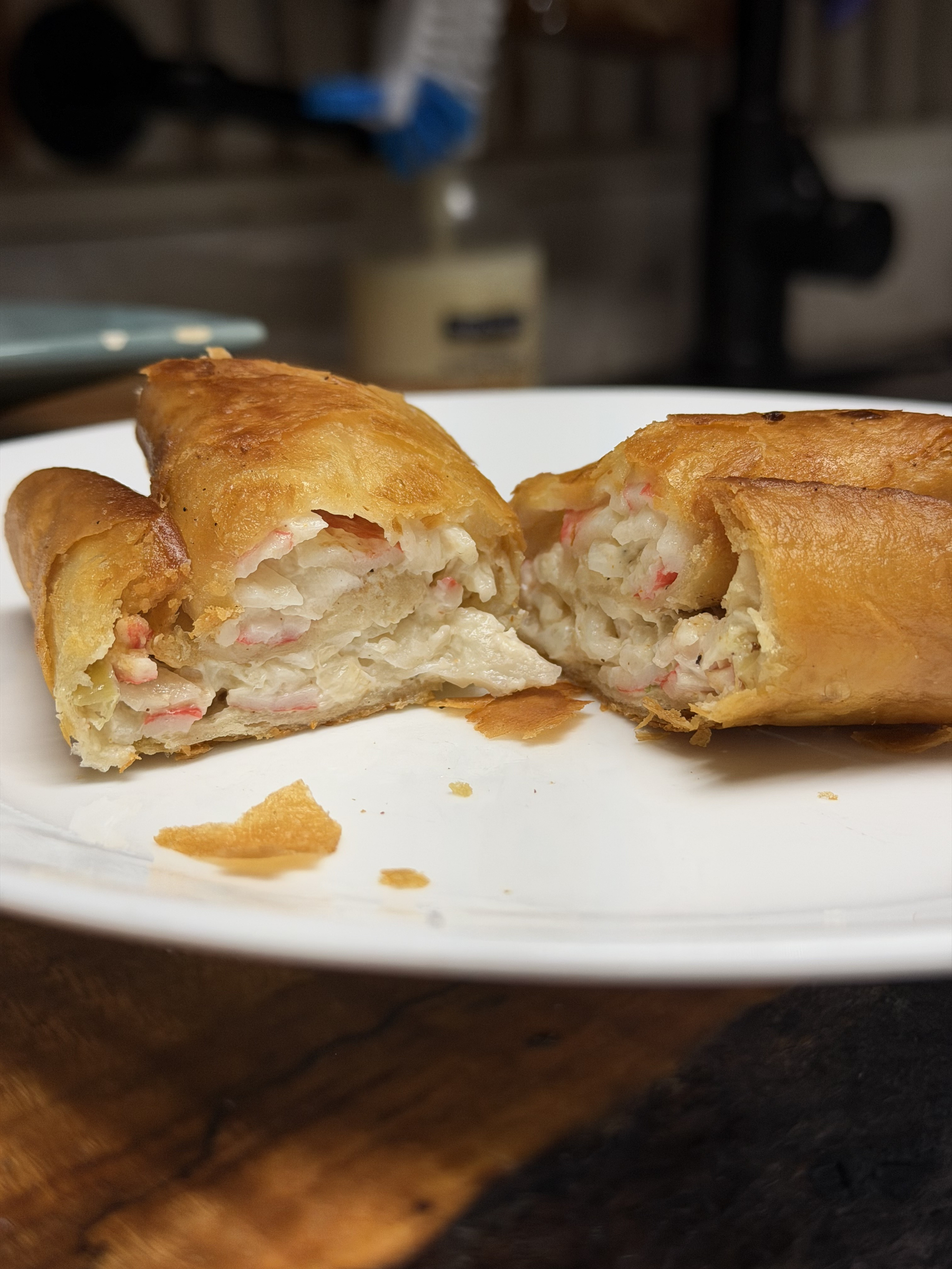 A crispy pastry split in half reveals a creamy filling of imitation crab mixed with diced green onion, served on a white plate with pastry crumbs scattered nearby.
